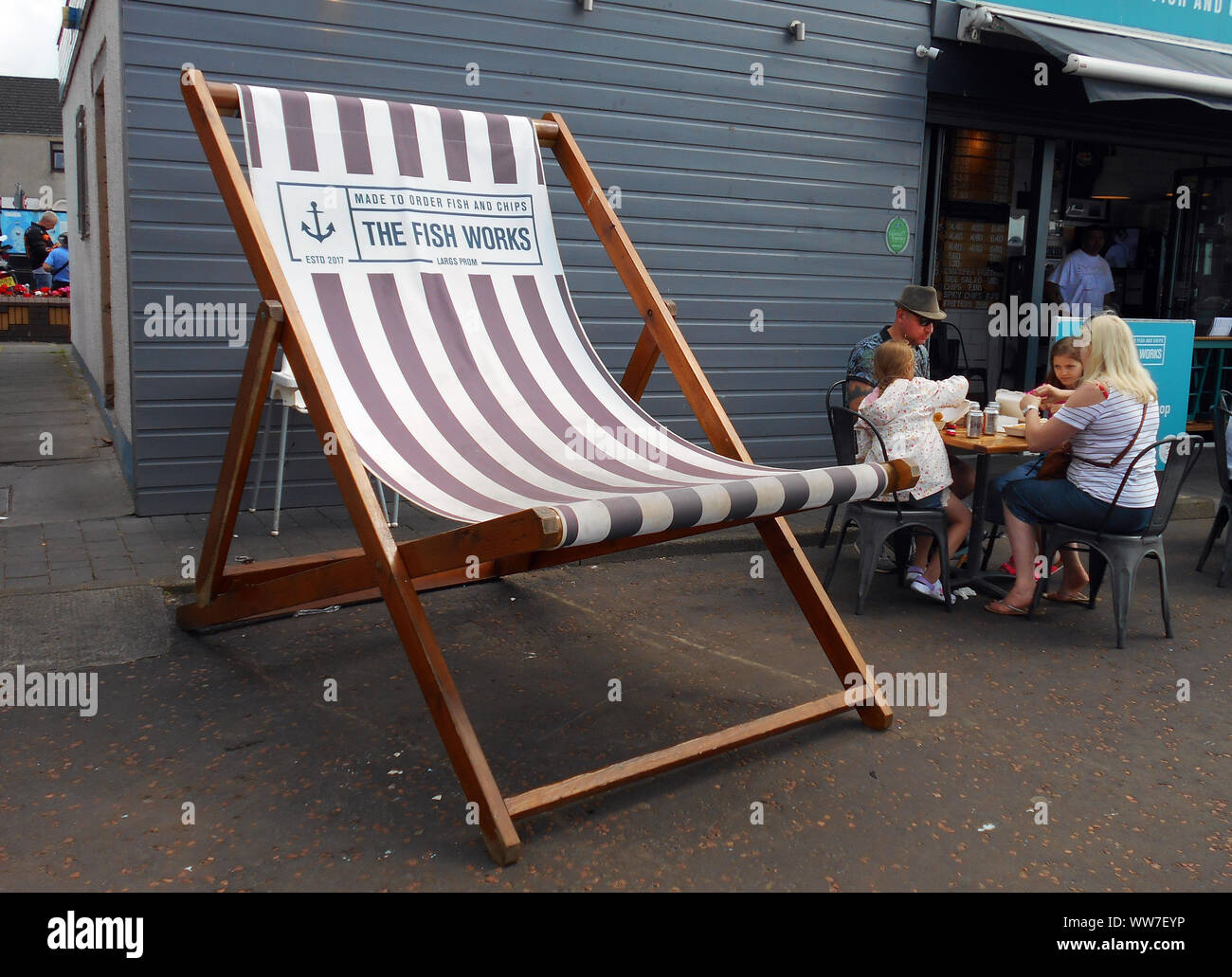 An extra large sized deck chair sits alongside the fish and chip shop that it is advertising, on the seafront of the sea side town of Largs in the Firth of Clyde, Scotland. Customers are seen to be tucking in, nearby this huge chair! Alan Wylie/ALAMY © Stock Photo