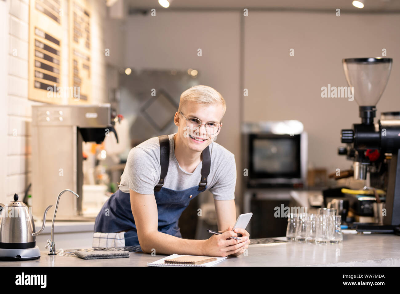 Young cheerful barista in workwear looking at you while bending over table Stock Photo