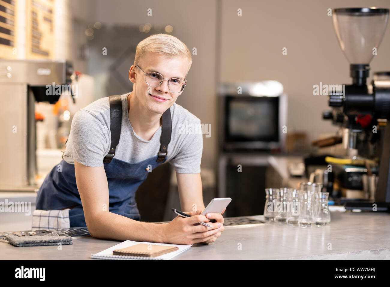 Happy waiter with pen and smartphone standing by workplace in front of camera Stock Photo