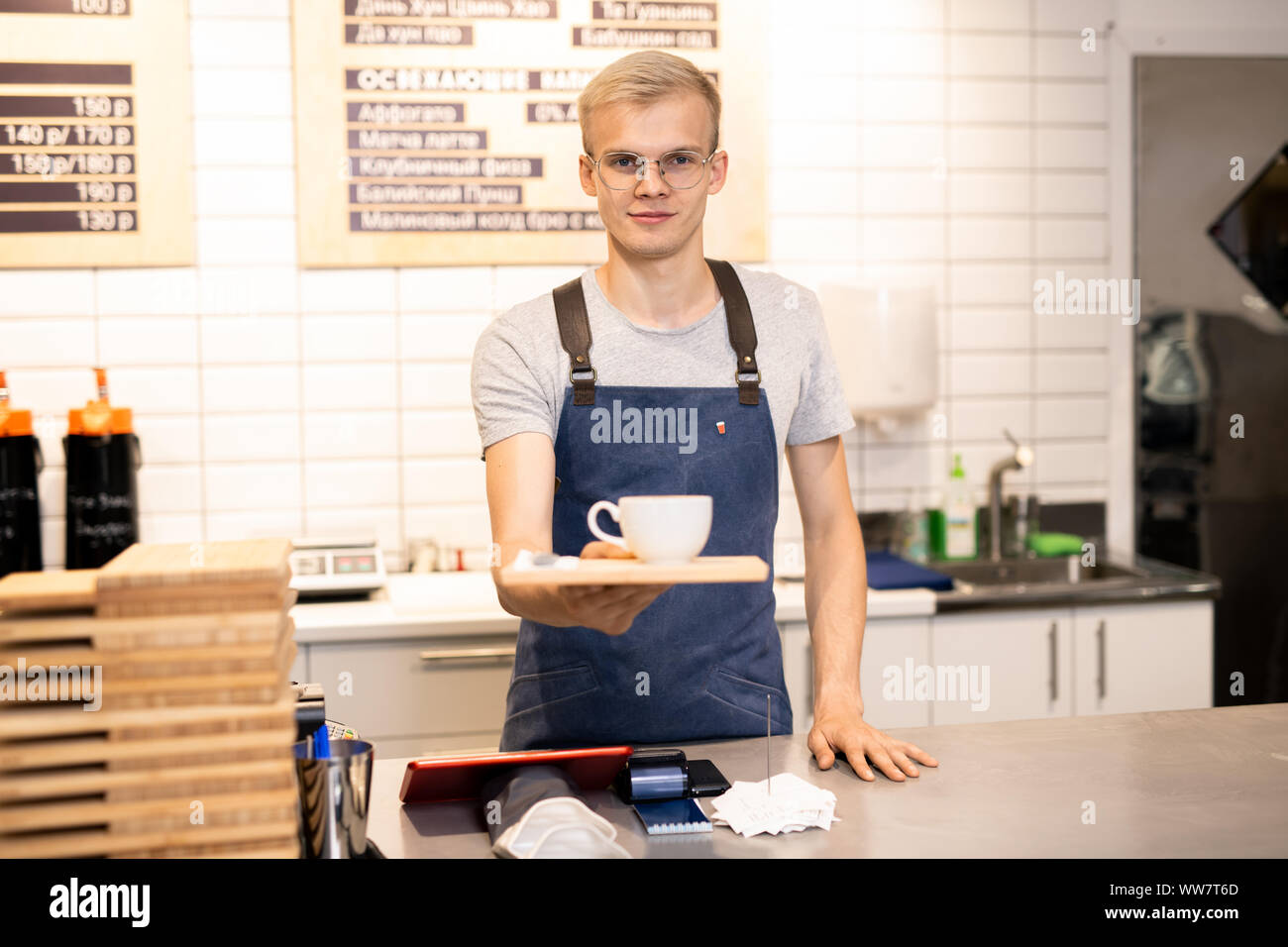 Young waiter in eyeglasses and workwear giving you tray with cup of coffee Stock Photo