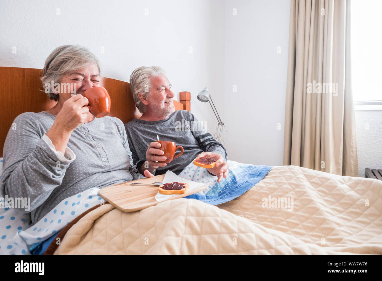 adult people aged couple having fun laughing and smiling on the bed at home. indoor bedroom scene family having brekfast lay down. window light and bright image Stock Photo