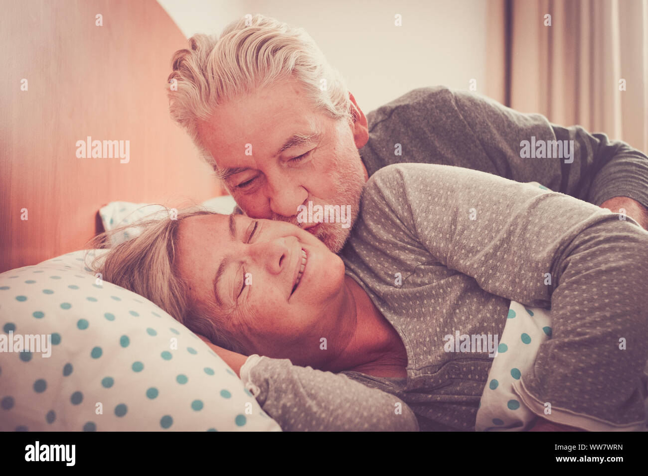 Couple of senior man and woman waking up and smiling with a hug while are in the bed at home. Vintage filter and light in the back. The man kiss the woman with love Stock Photo