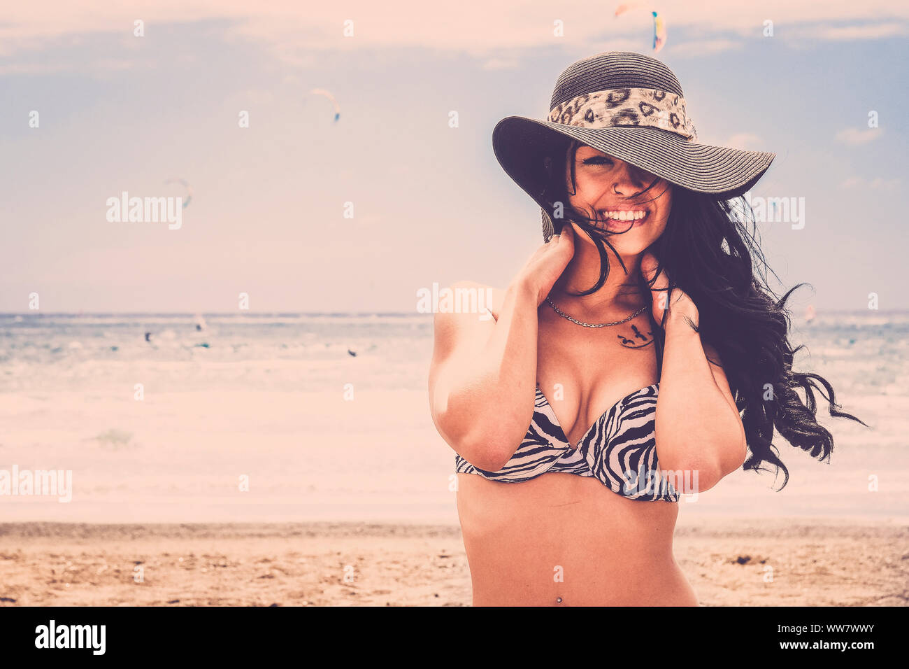 young smile beautiful woman smile at te camera on the beach in Tenerife. Long black hair touching hre shoulder back. Big grey hat and kitesurfers on the back. Stock Photo
