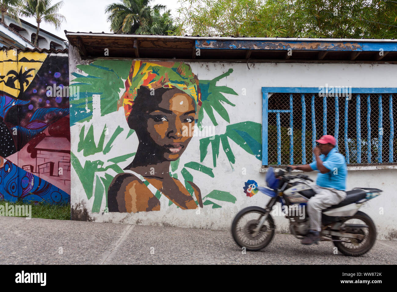 Man on motorcycle driving in front of wall of a house with coloured graffiti of a woman Stock Photo