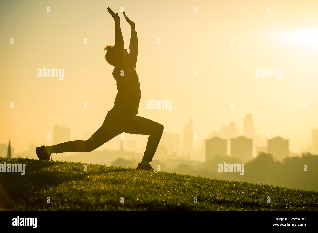 Sunrise silhouette of a man stretching in a relaxed Anjaneyasana yoga lunge on the grassy top of a hill front of a misty view of the London skyline Stock Photo