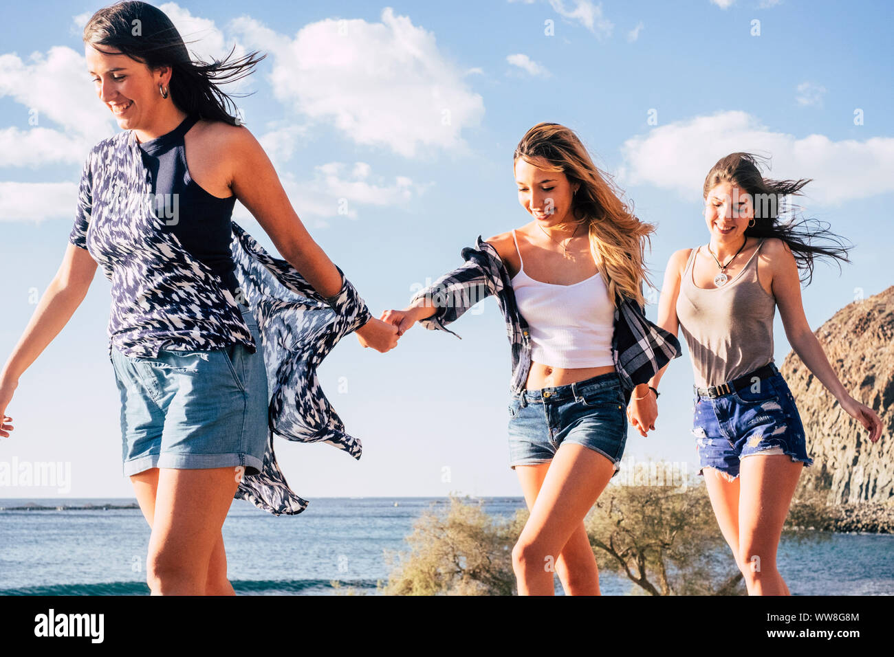 three young caucasian pretty girls walking together taking hands each other smiling and laughing, cheerful concept of happy women in outdoor leisure activity near the ocean and mountains Stock Photo