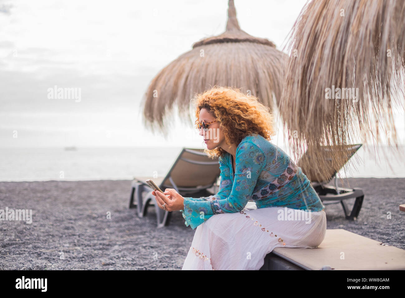 alternative office at the beach with ocean in background for a beautiful middle age woman working on her smartphone sitting on a seats under the umbrella sun, vacation lonely lady connecting with friends Stock Photo