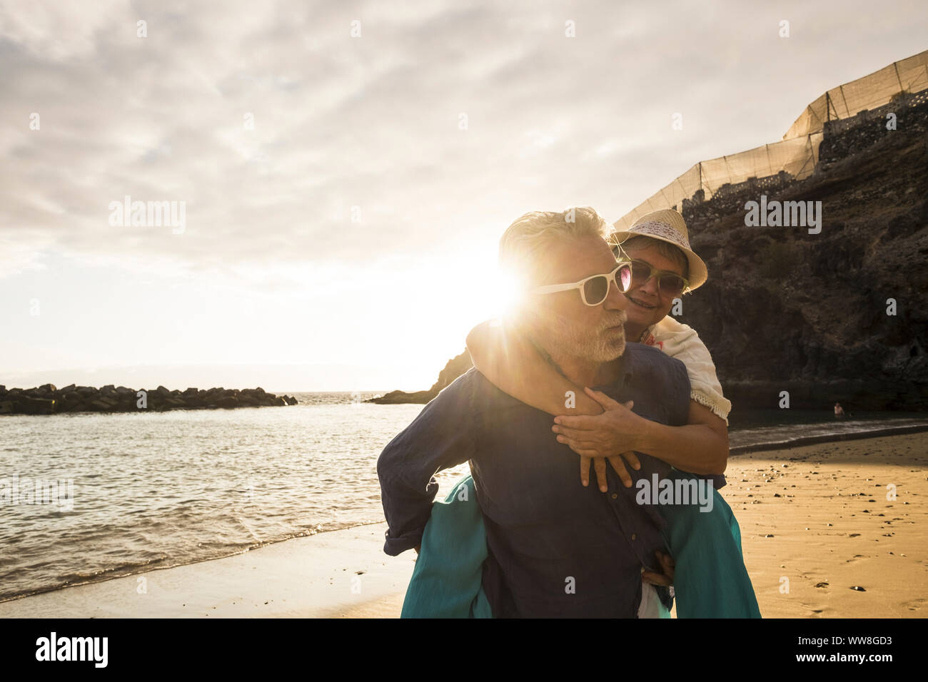 Happy senior adult caucasian couple in love, the man carry on his back the beautiful woman and both smiles, affective and together forever life concept in vacation time with sunset backlight and ocean waves Stock Photo