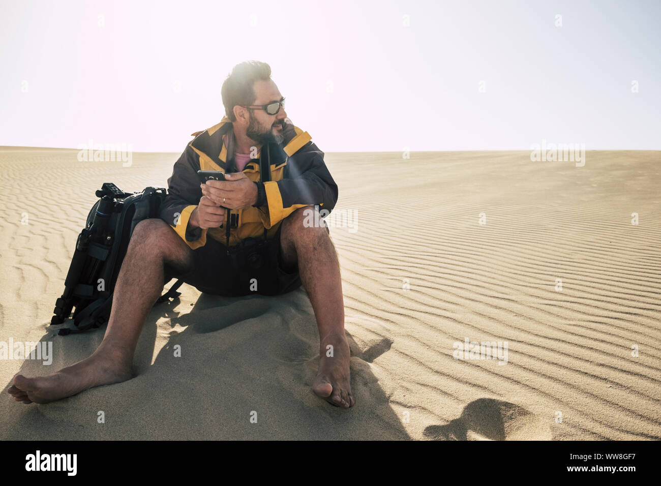 lonely caucasian middle age man travel barefoot in the desert with backpack and phone and tripod to take pictures, alternative travel vacation concept to enjoy freedom and emotions and feel the real nature in the world Stock Photo