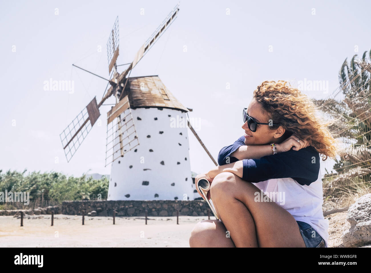 nice curly lady sitting and relaxing with wind in the hair and a wind mill on the background, country side scenic place for peace and relaxation concept for young people that travel in wanderlust and enjoy the world Stock Photo
