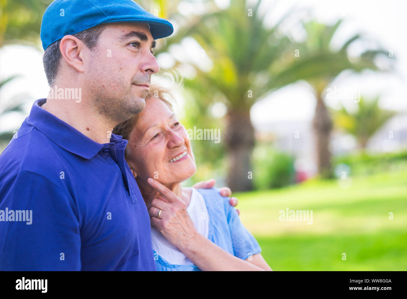 different ages couple like mother and son stay hugged together in outdoor leisure activity with love and emotions, enjoy the day in the park with green plants tropical background, bright colors Stock Photo