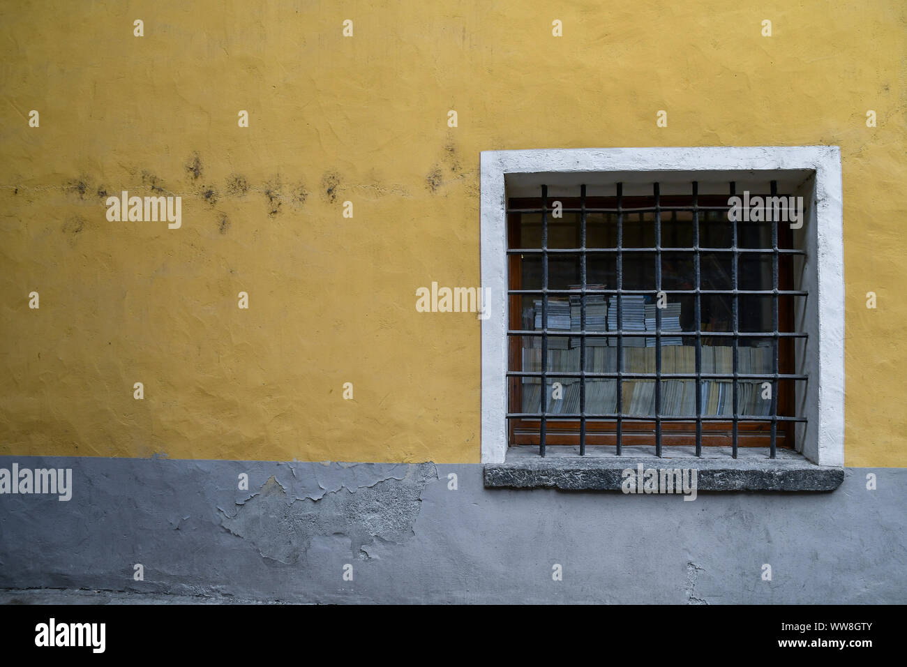 Close-up of the small window of an old building with iron bars, stacks of books behind the glass and yellow and gray wall, Aosta, Aosta Valley, Italy Stock Photo