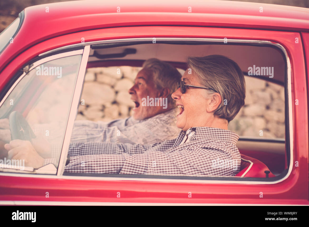 Elderly senior couple with hat, with glasses, with gray and white hair, with casual shirt, on vintage red car on vacation enjoying time and life. With a cheerful mobile phone smiling and going crazy together Stock Photo