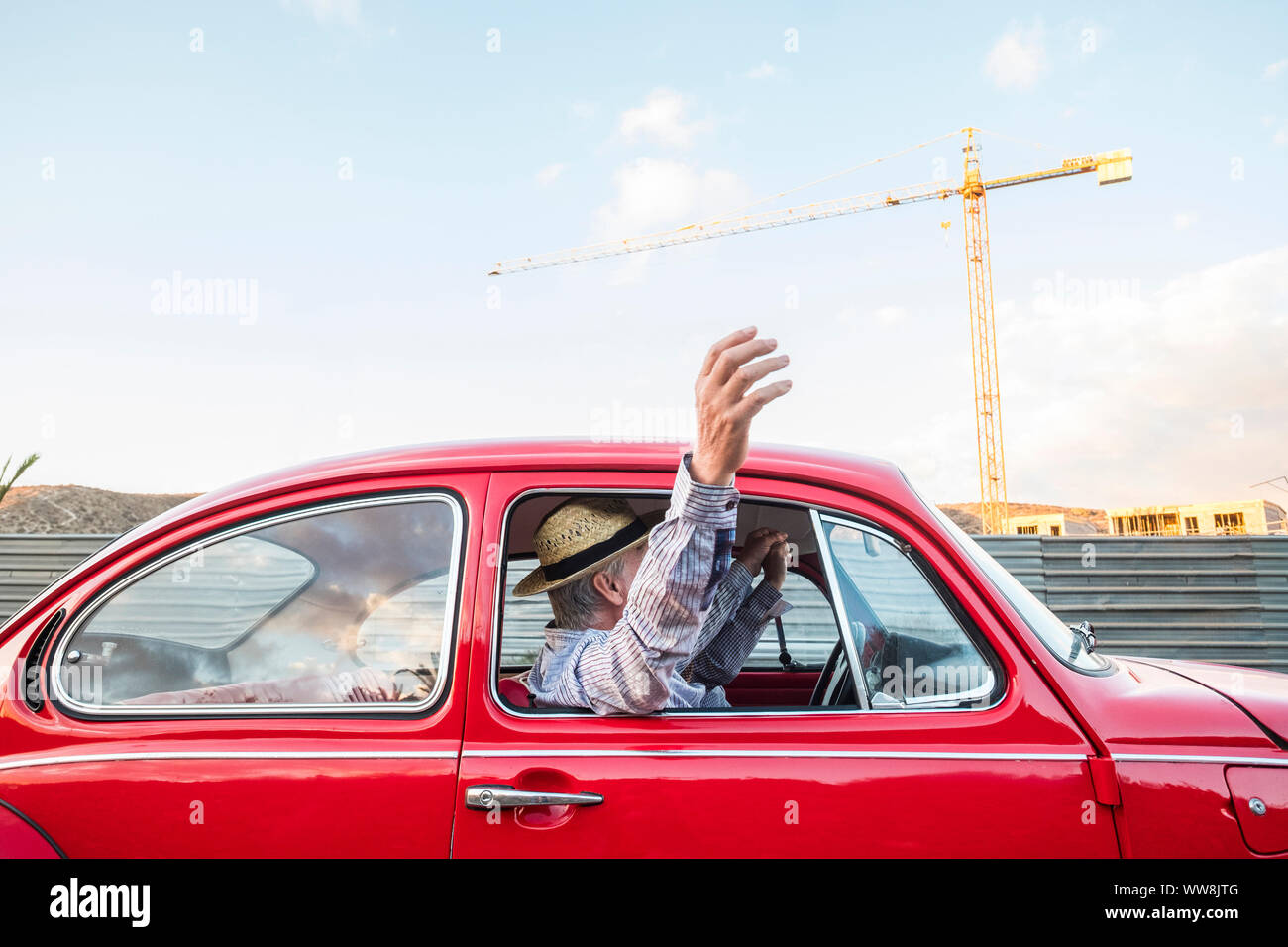 caucasian mature couple travel and drive on a red vintage car. vacation and wonderlust concpet for happy people moving around the world. home building in the background Stock Photo