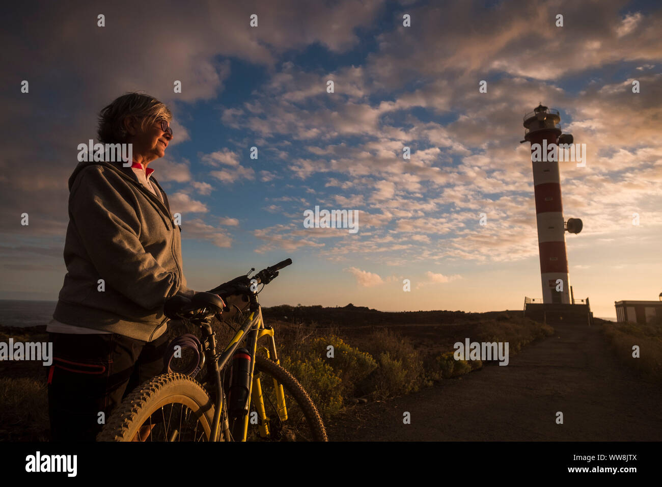 adult senior beautiful caucasian woman biker standing and enjoy the sunset near the coast and the ocean. mountain bikes sport. lighthouse in the backgorund and amazing scenic colored place Stock Photo