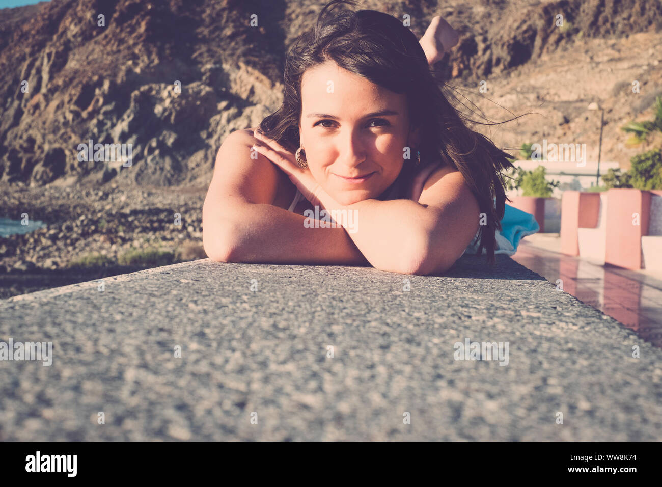 long hair spanish young woman erlaxing lay down on a wall near the beach. Sunlight on her beauty face, smile and enjoy the time. Stock Photo