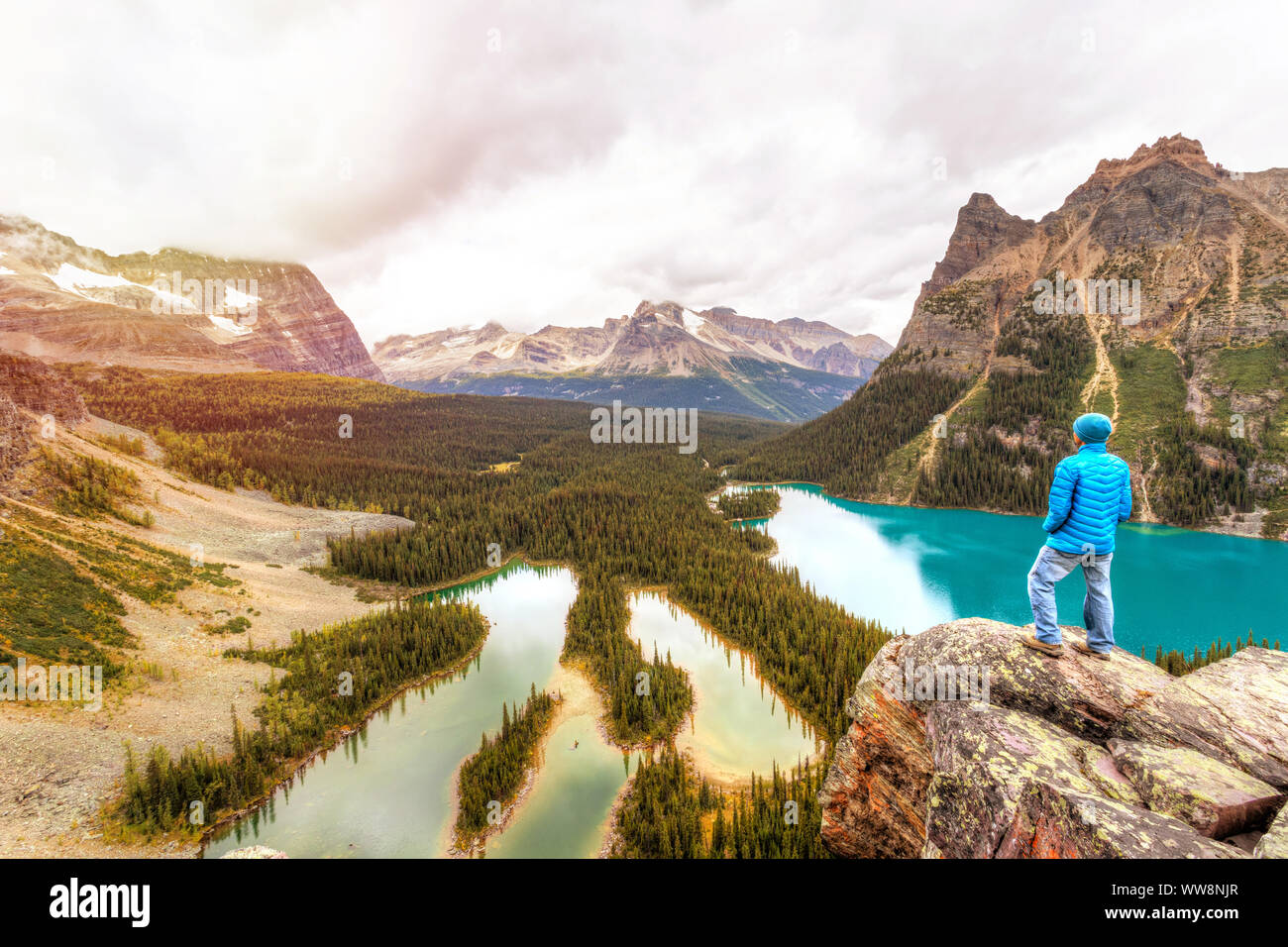 Sunset over Lake O'Hara as hiker stands atop Opabin Prospect overlooking Lake O'Hara and Mary Lake with the Canadian Rockies in the background surroun Stock Photo