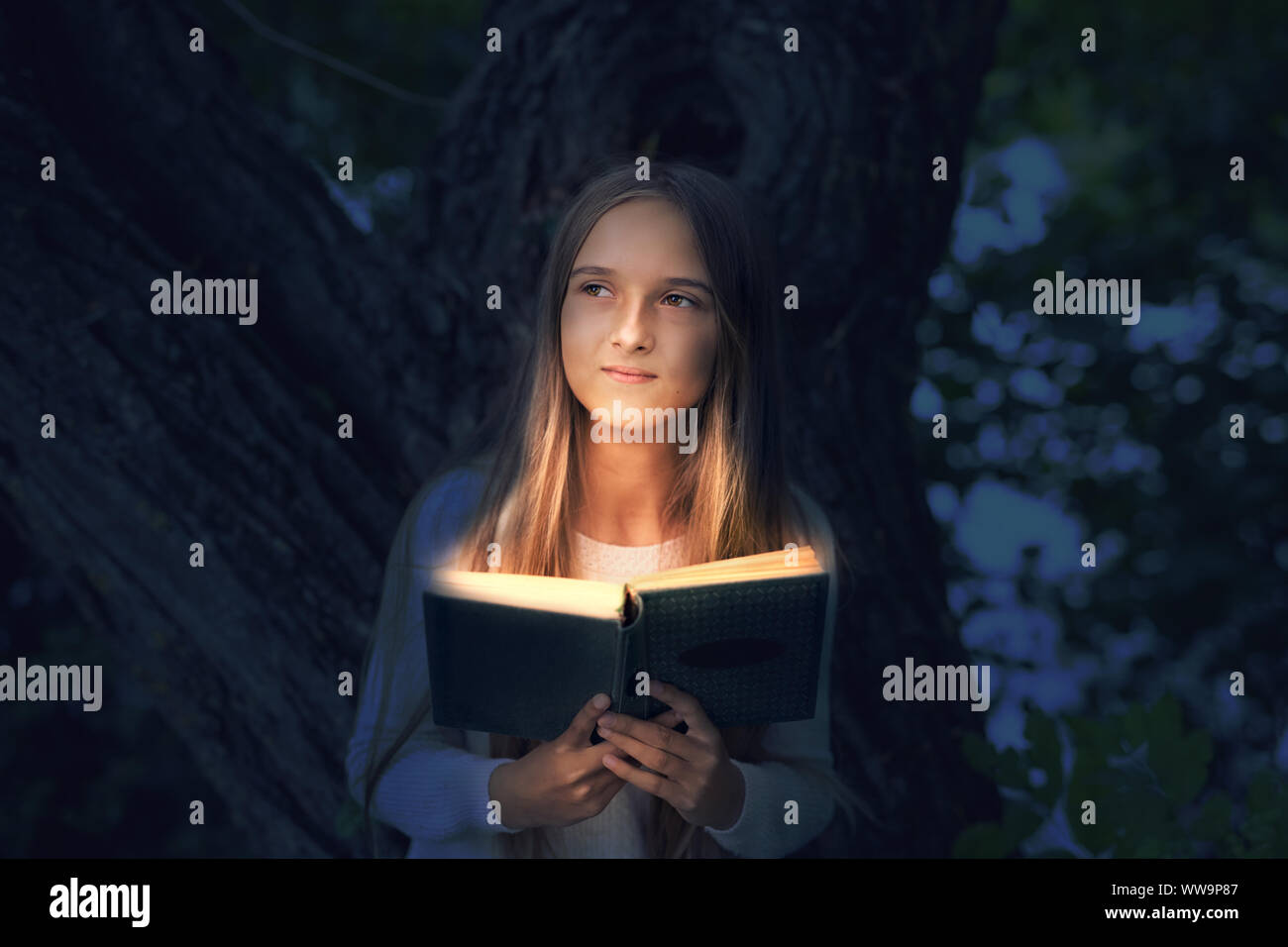 A cute teenage girl with long blonde hair is reading a magic book with light rays coming out from the inside, in a forest near a tree on a dark colore Stock Photo