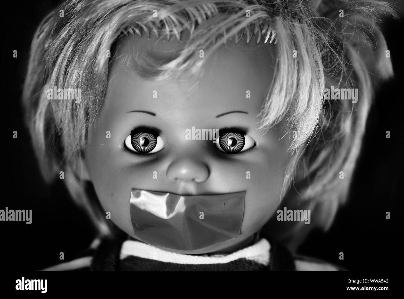 Creepy doll's head in black and white. A scary face of a weird vintage doll with its mouth taped on black background Stock Photo