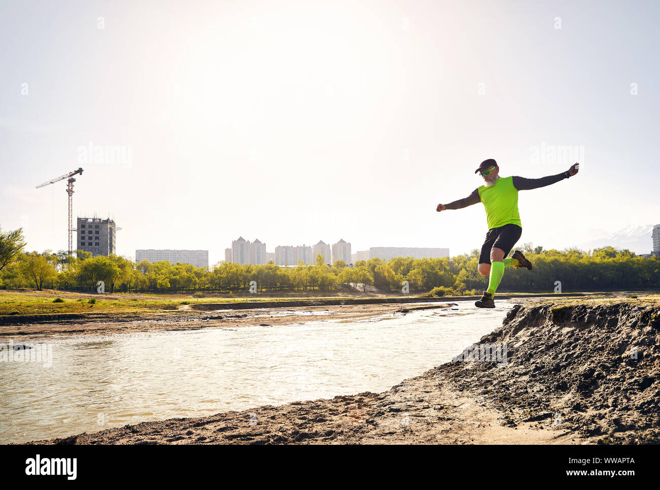 Elderly man running in park near the river with buildings and mountain background in the morning. Healthy lifestyle concept Stock Photo