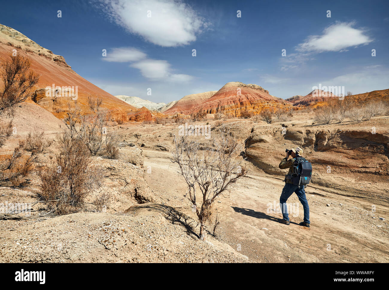 Tourist with backpack and camera taking a picture at the dusty canyon on surreal red mountains against blue sky in the desert Stock Photo