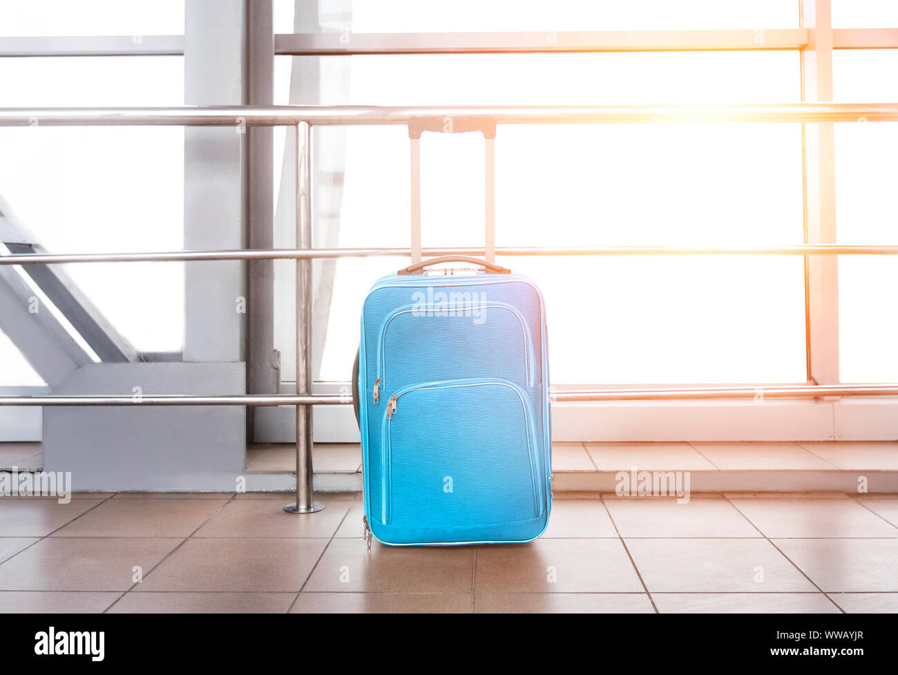 Blue suitcase at airport building, against window with sun flare Stock