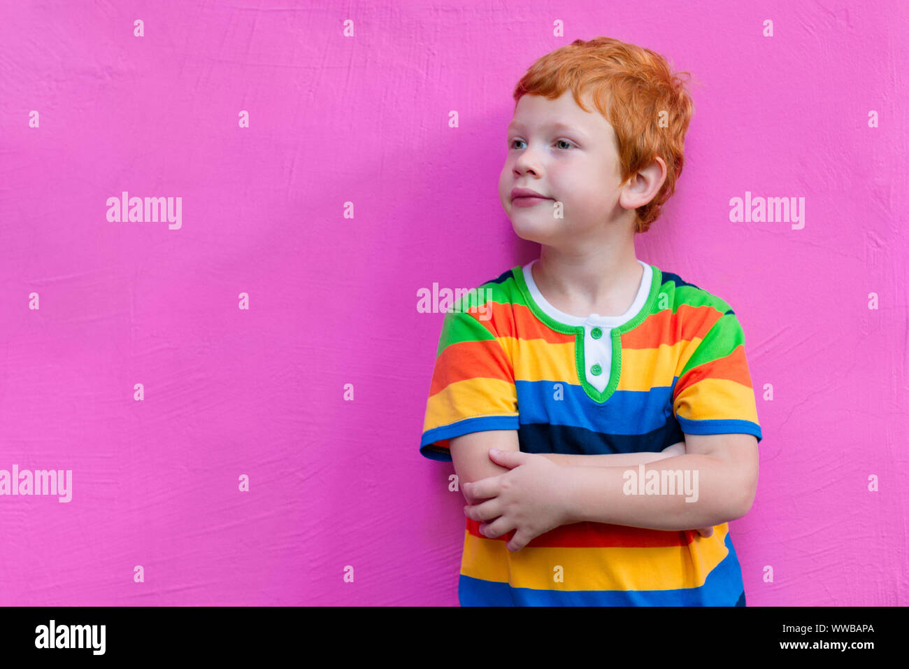 Fashionable little boy. Red-haired handsome kid model wearing trendy striped casual shirt posing against pink wall background enjoying good summer Stock Photo