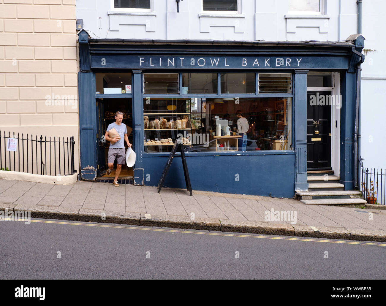 Person coming out of Flint owl Bakery on inclined High Street, Lewes, carrying bread purchase from establishment. Lewes, High Street Stock Photo