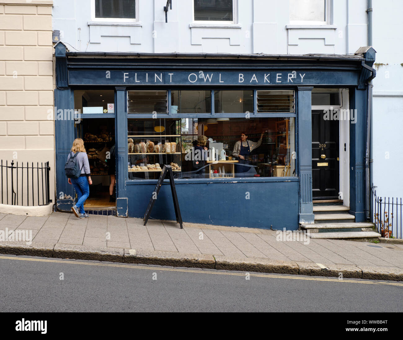 Person walking into  Flint owl Bakery on inclined High Street, Lewes, while through window can see barista taking coffee order from customer. Stock Photo