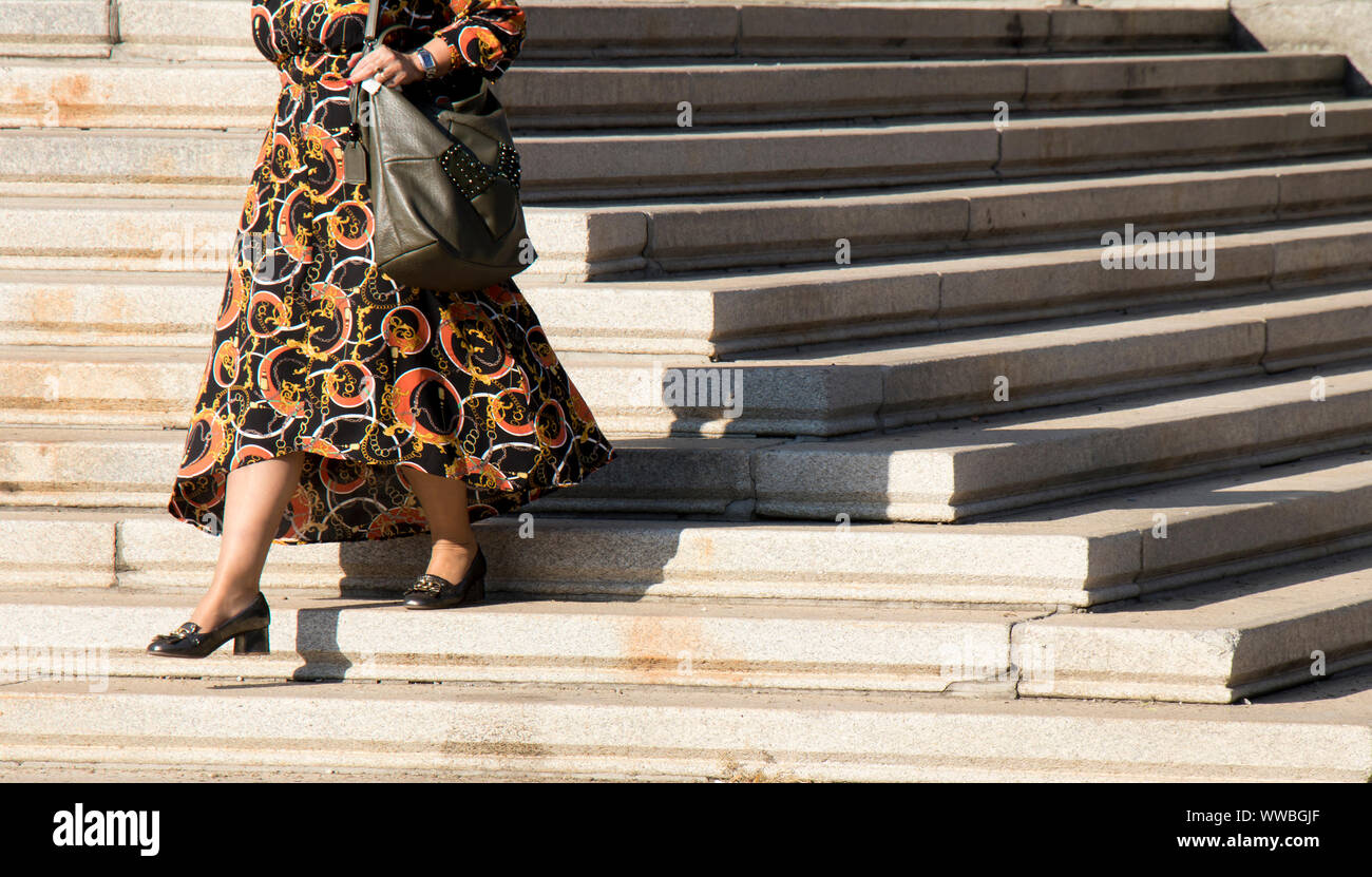 Legs of a mature plus size woman walking down city stairs in  long black and orange dress on a sunny day Stock Photo