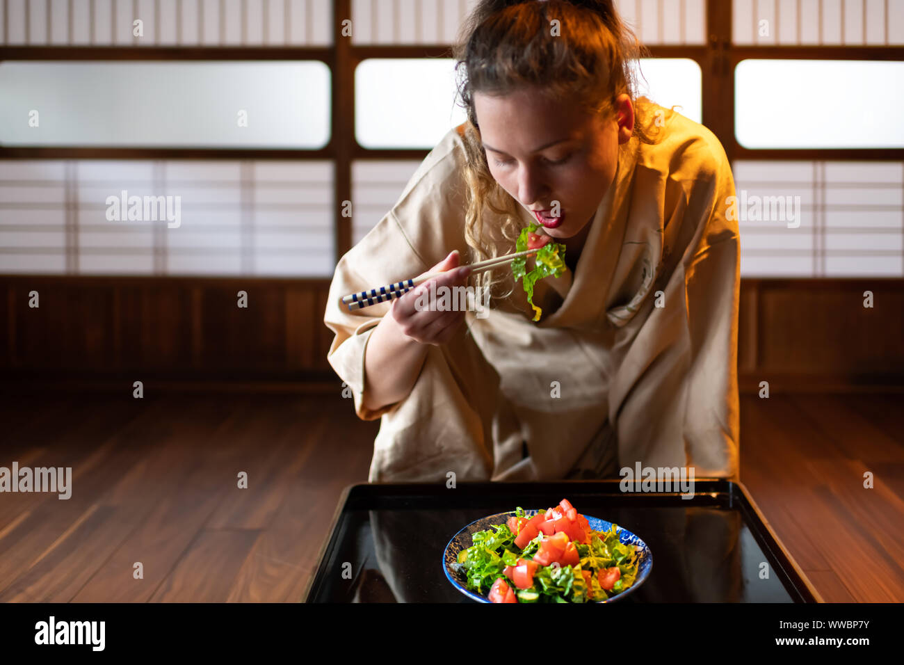 Young woman in kimono seiza sitting at traditional Japanese house or ryokan room by table holding chopsticks, eating salad from plate by shoji sliding Stock Photo