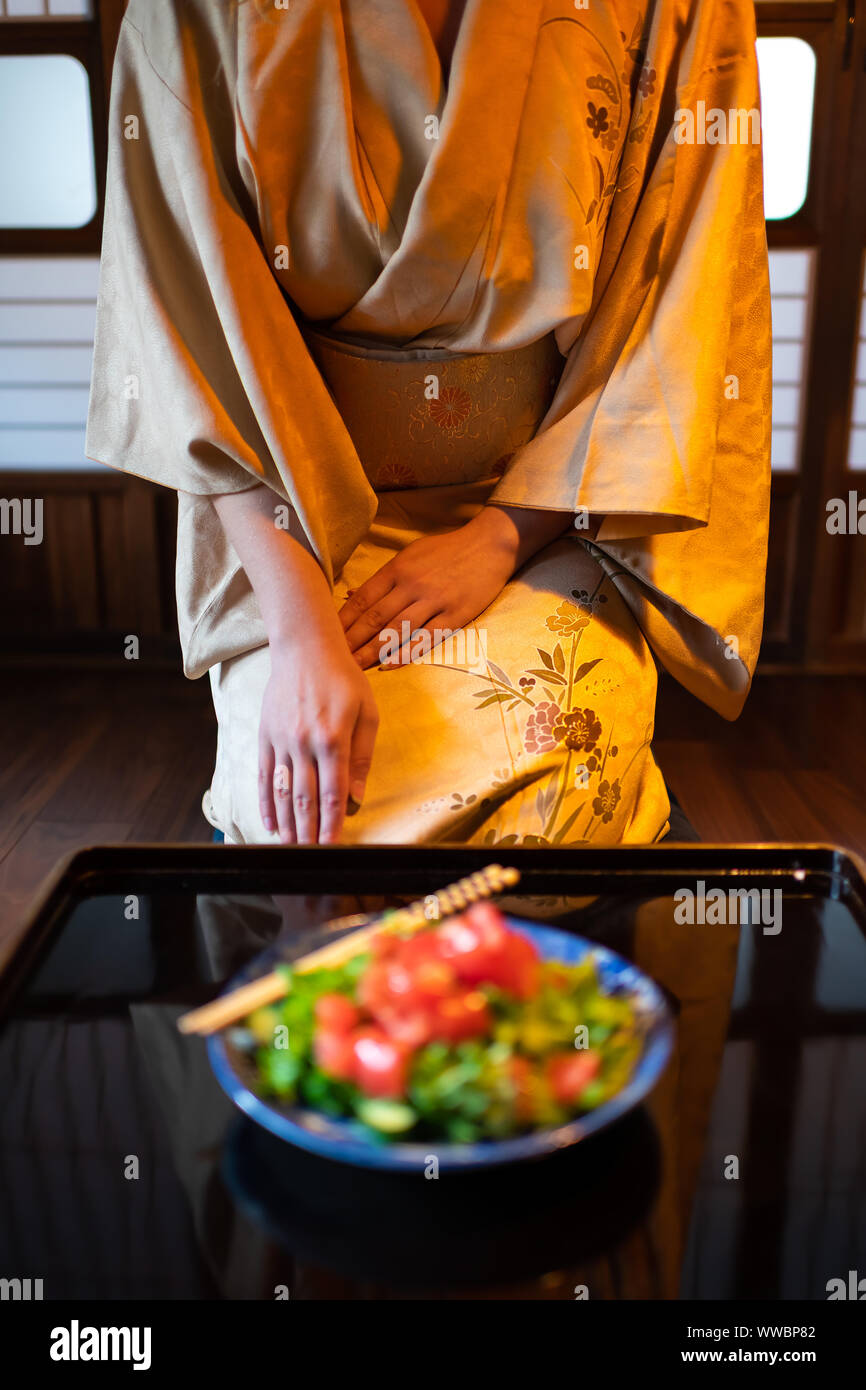 Young woman in kimono, obi seiza sitting at traditional Japanese house or ryokan restaurant room by table with salad plates and chopsticks, shoji slid Stock Photo