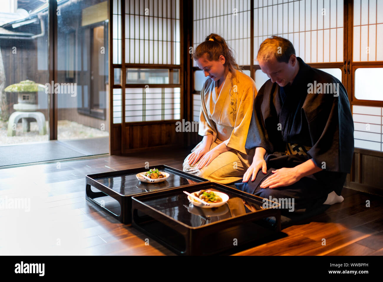 Couple in kimono bowing ojigi, seiza sitting at traditional Japanese home room by food plates on table with shoji sliding paper doors looking garden w Stock Photo