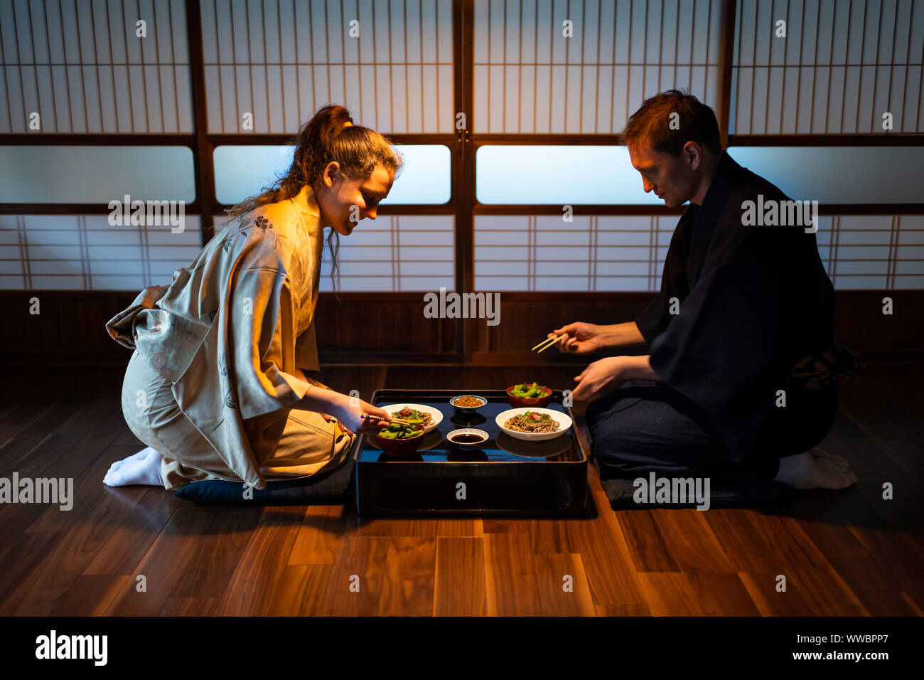 Young couple seiza sitting on floor pillows with Japanese ryokan kaiseki dishes, eating soba noodles, natto, edamame and soy sauce on table by sliding Stock Photo