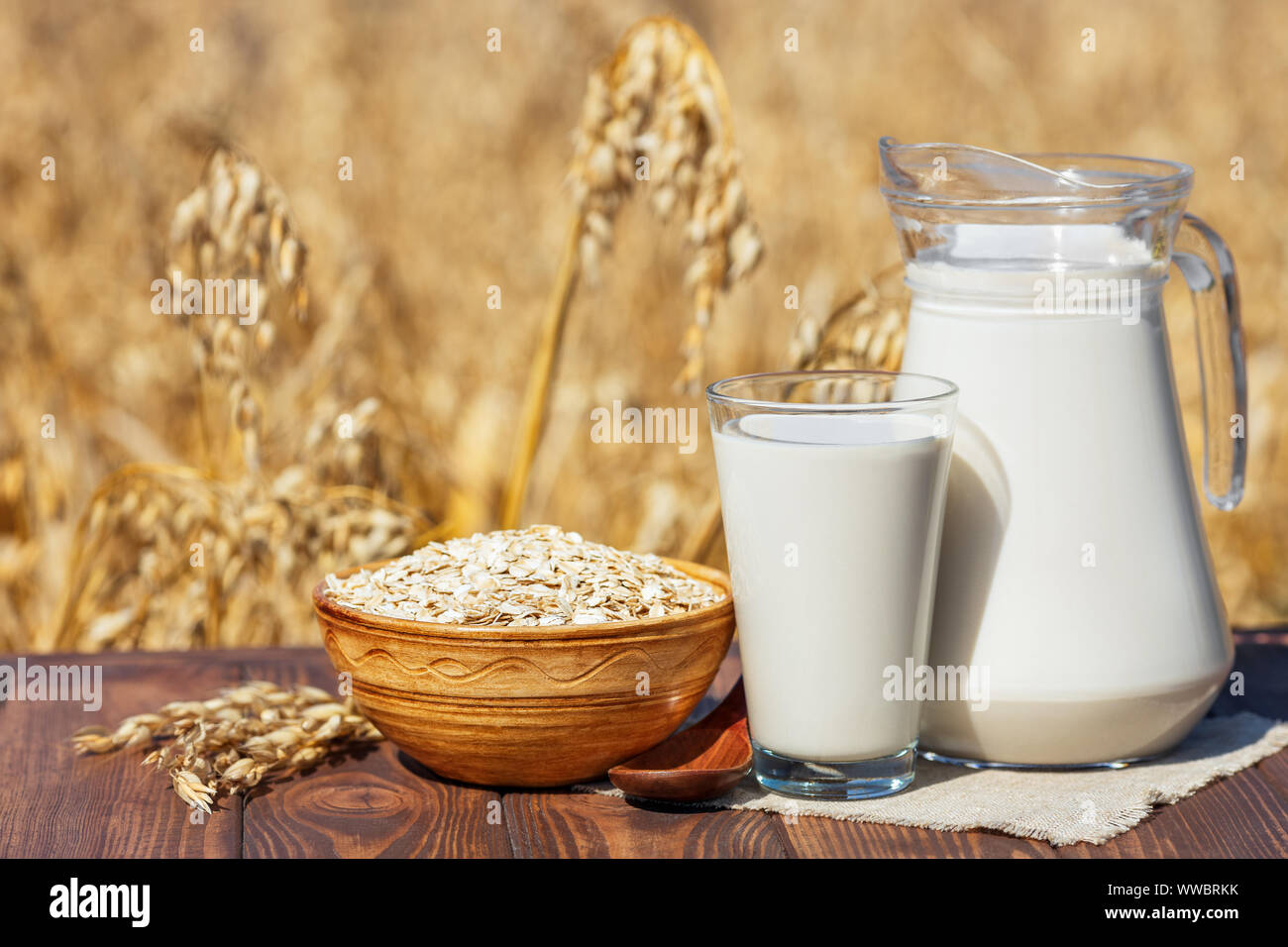vegan oat milk in glass and jug with uncooked oatmeal in bowl on table over against ripe cereal field Stock Photo