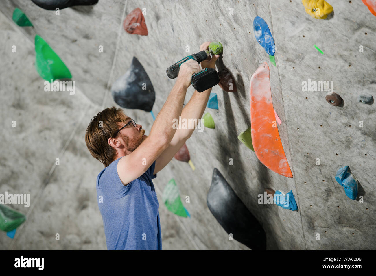 active young bearded man in glasses building climbing wall indoors using drill tool Stock Photo