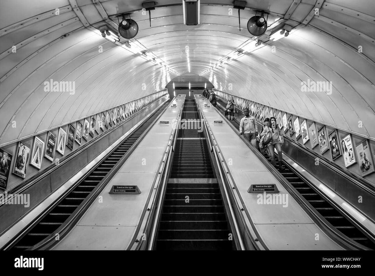 Monochrome view looking up moving escalators inside London Underground Tube station. Black & white front view of passengers travelling down escalator. Stock Photo