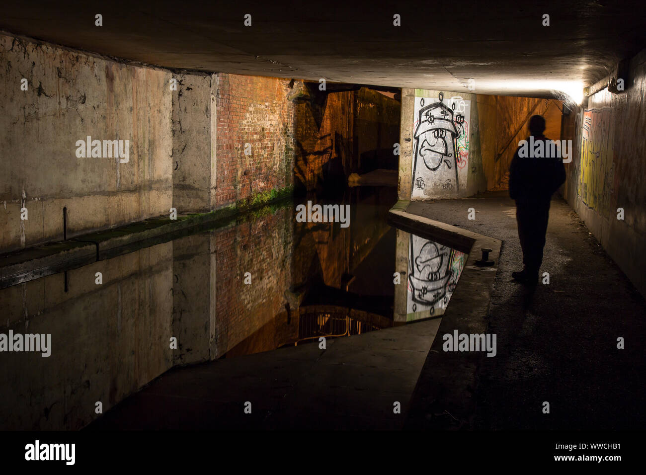 Dark, moody, solitary figure standing by an urban canal in UK pedestrian underpass at night, staring at urban graffiti artwork sprayed on walls Stock Photo