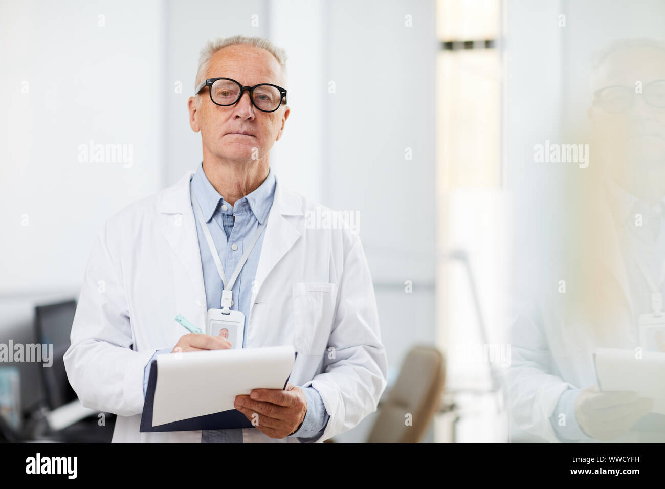 Waist up portrait of senior doctor holding clipboard and looking away pensively while taking notes standing in office, copy space Stock Photo