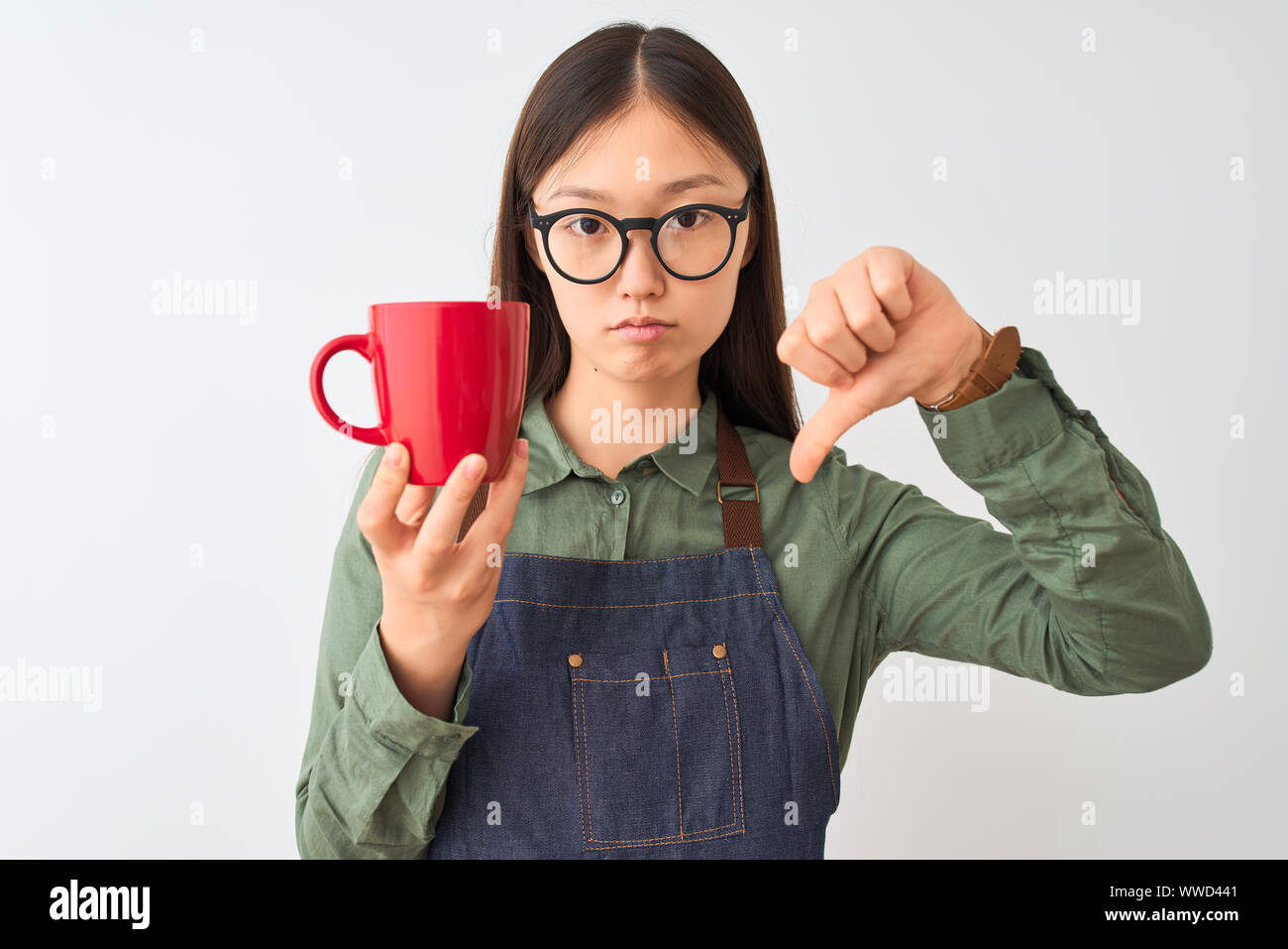 Chinese barista woman wearing apron glasses drinking coffee over isolated white background with angry face, negative sign showing dislike with thumbs Stock Photo