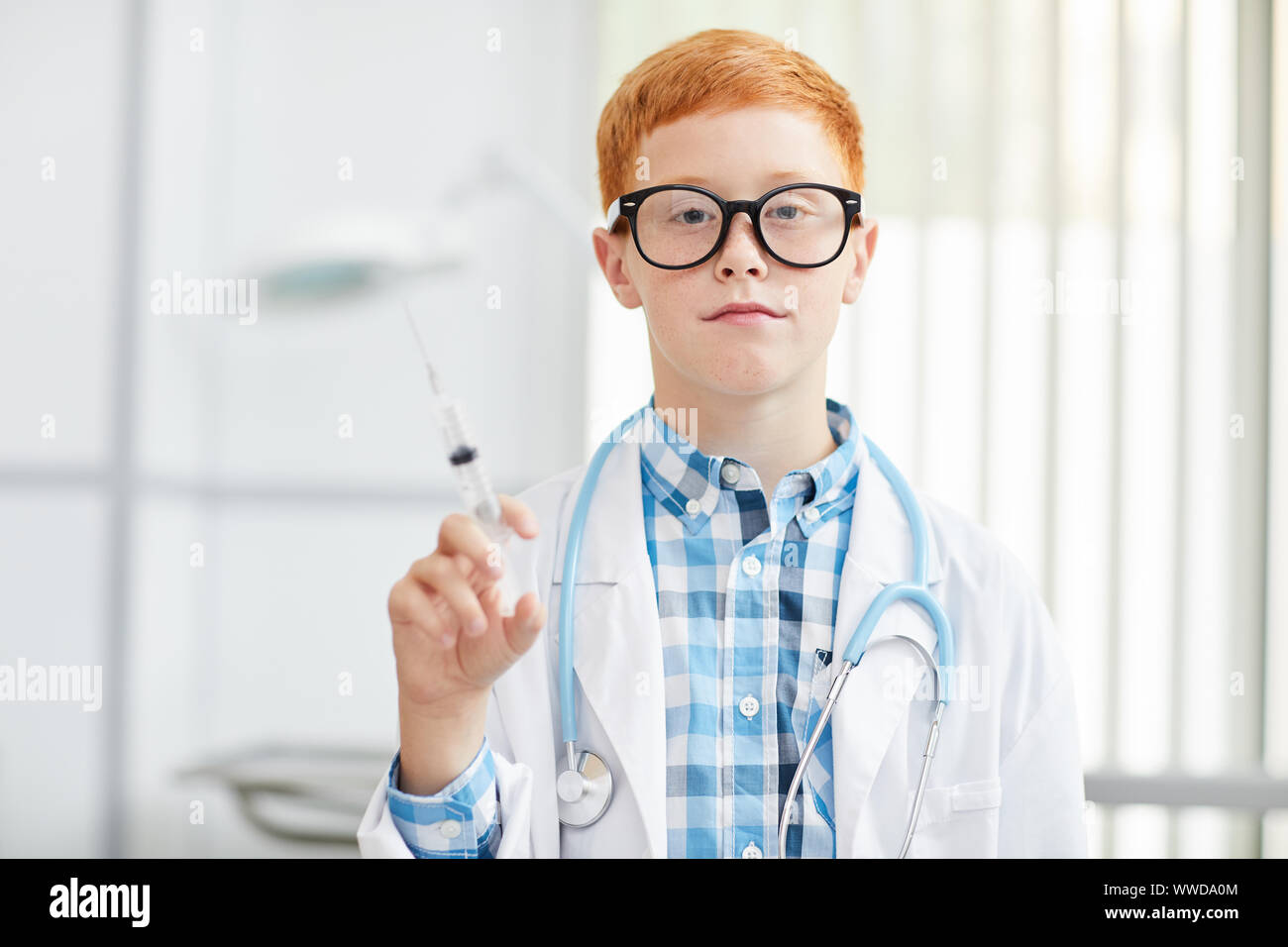 Waist up portrait of cute little boy wearing lab coat posing as doctor holding syringe and looking at camera, copy space Stock Photo