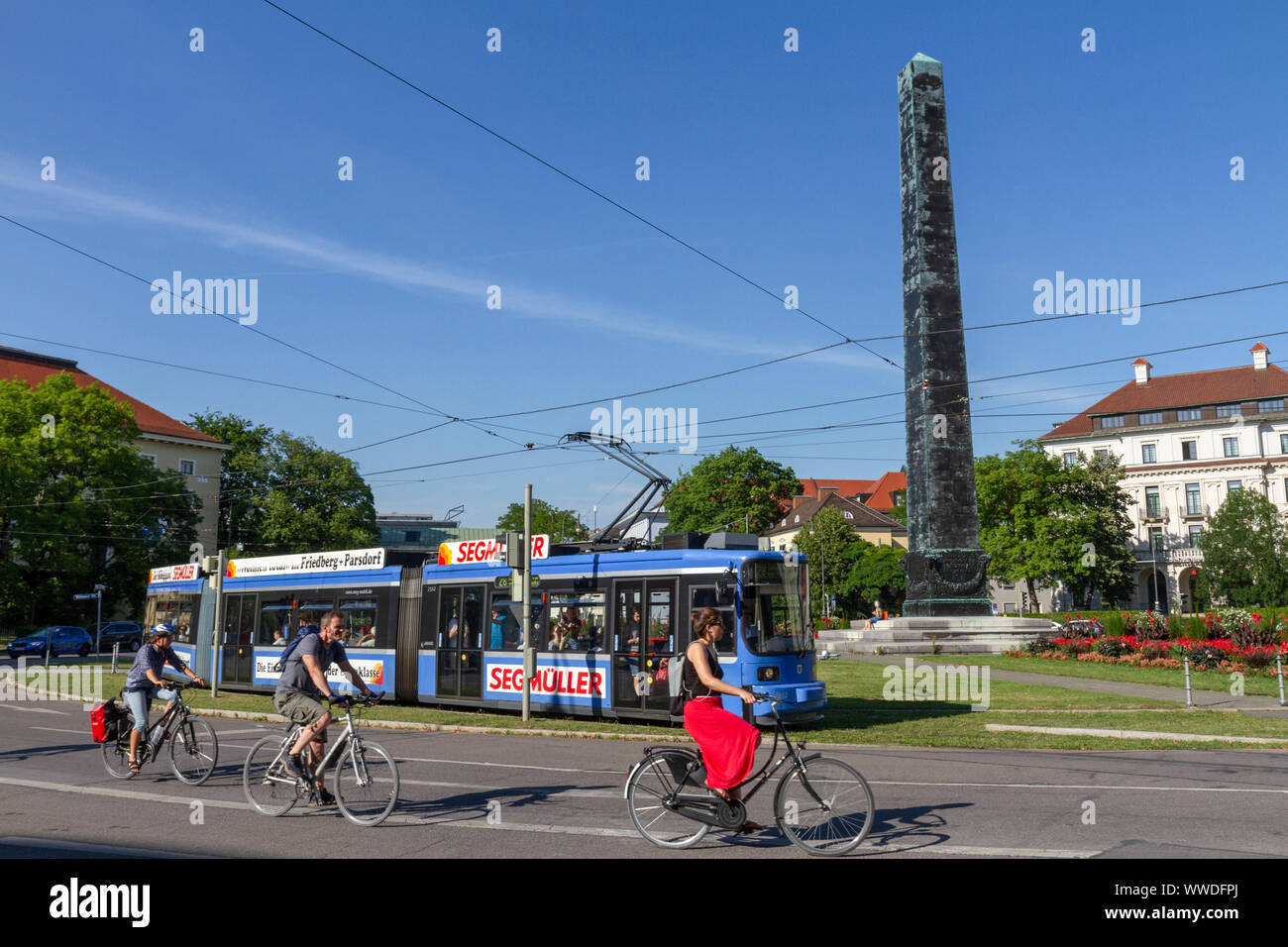 Cyclists and Munich electric tram passing the Mariensäule, a Marian column in Karolinenplatz (Caroline’s Plaza), Munich, Bavaria, Germany. Stock Photo