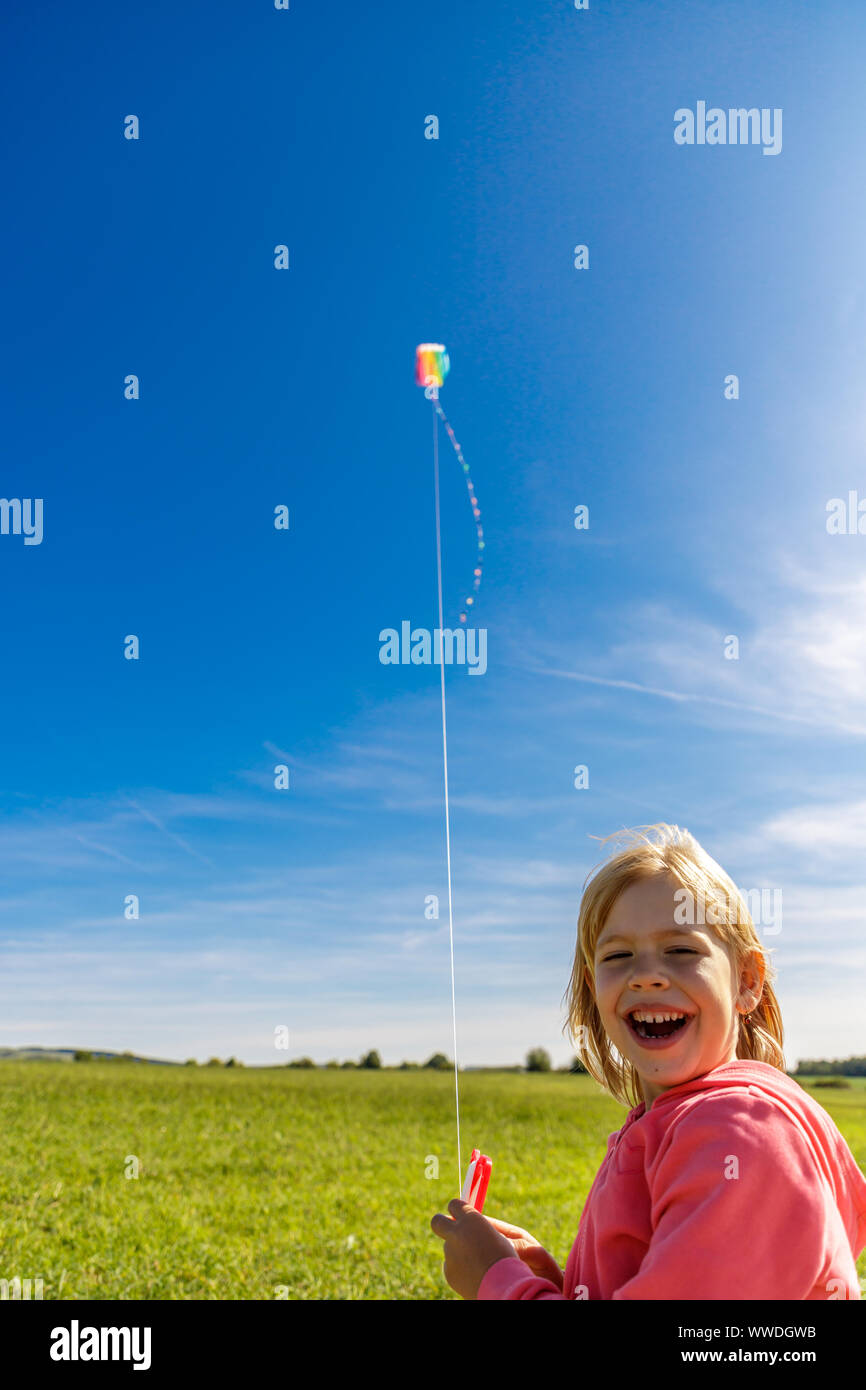 Pretty laughing little girl with blond hair on field in pink sweatshirt looks at colorful dragon in blue sky with soft clouds Stock Photo