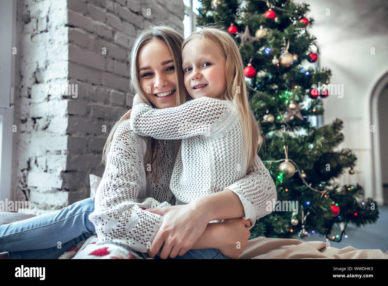 Happy young woman with cute daughter sitting on bed at home, enjoying family, traditional Christmas decoration, New Year holidays concept Stock Photo
