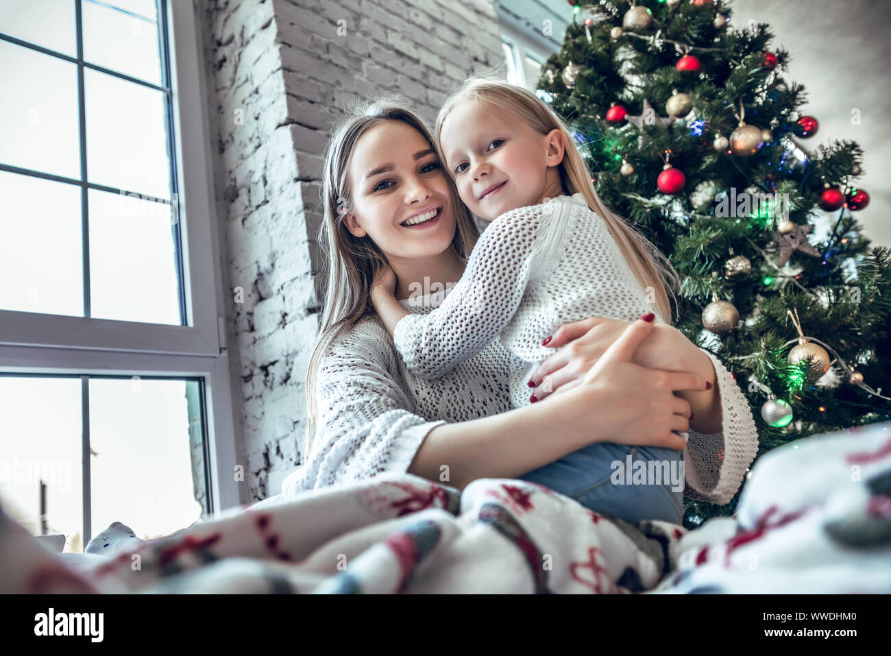 Cheerful mother and daughter sitting on the bed near beautiful decorated Christmas tree, spending New Year eve at home together, happy family concept Stock Photo