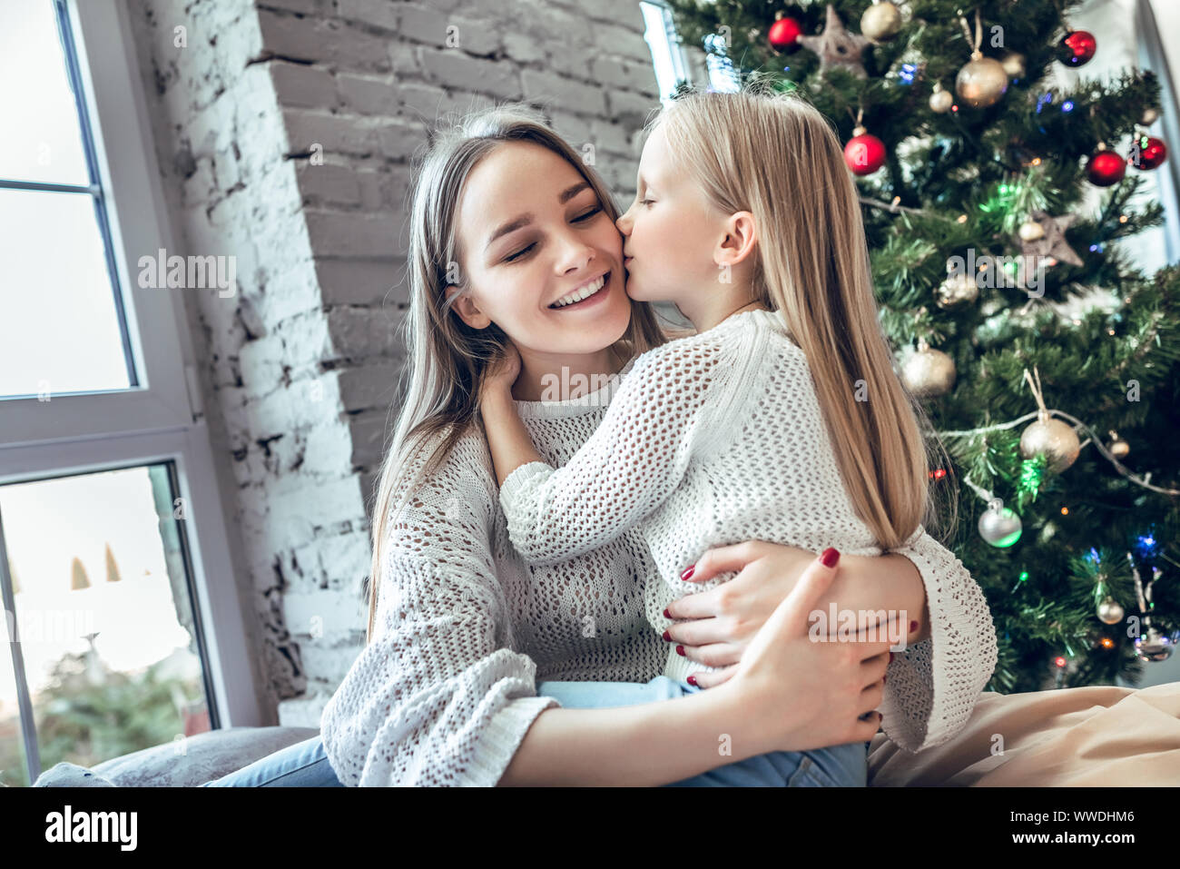 Little girl kissing her mom on the cheek on xmas tree background. spending New Year eve at home together, happy family concept Stock Photo