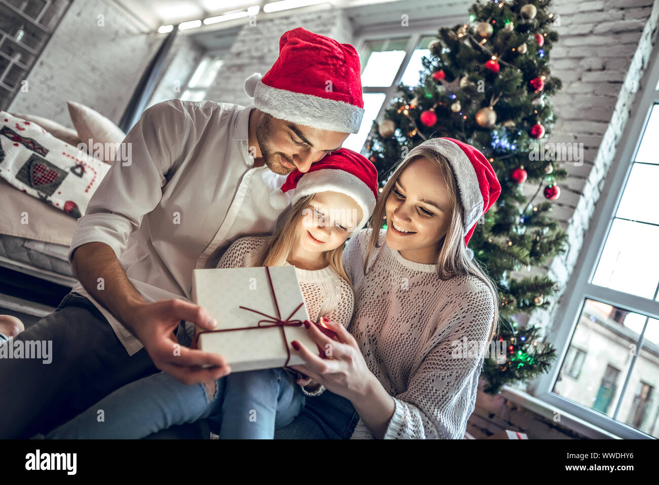 Young mother, father and little daughters opening a magical Christmas gift by a Christmas tree in cozy living room in winter Stock Photo