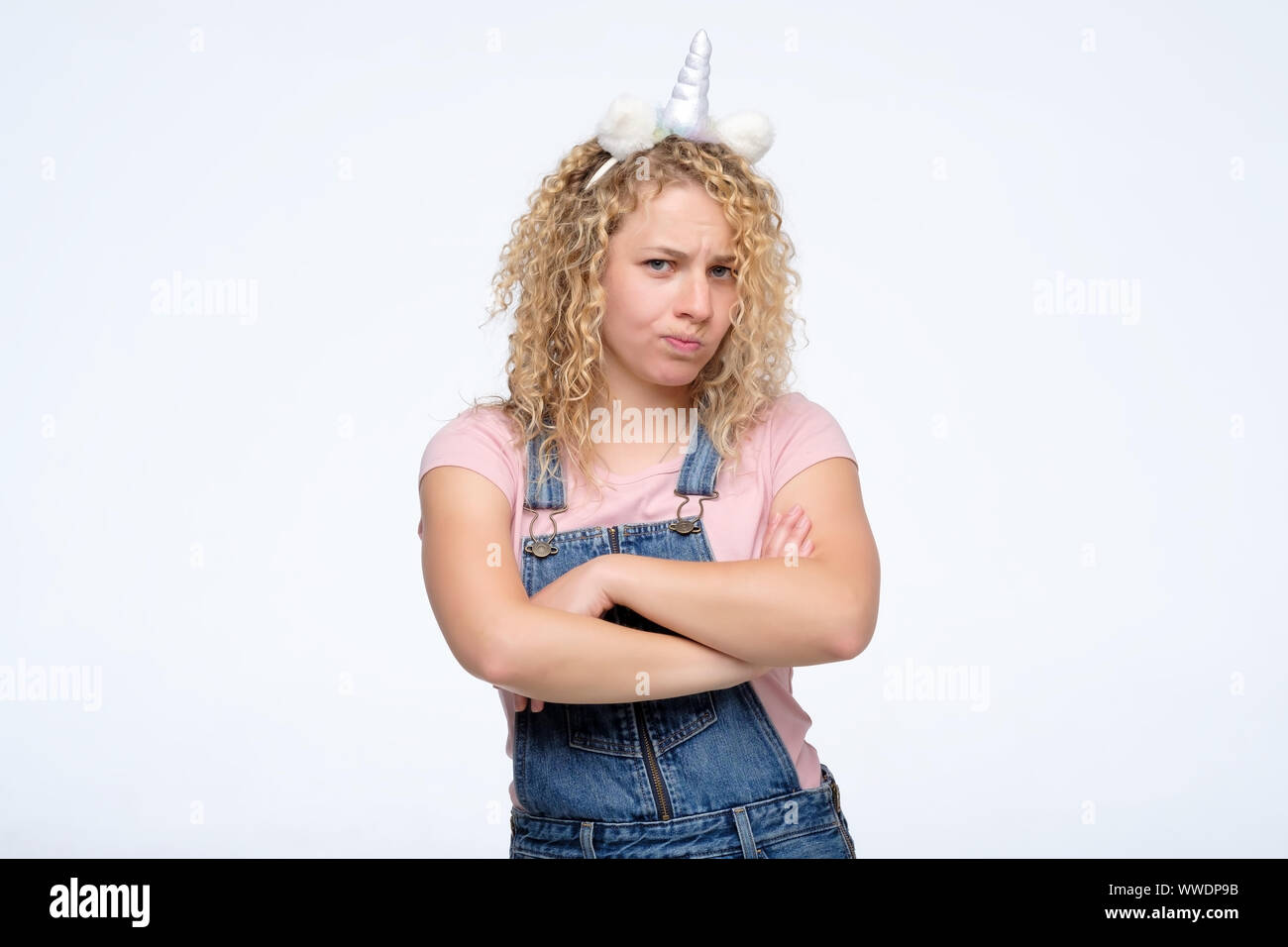 Young angry woman with curly hair and unicorn horn folded her arms, isolated studio shot. Negative facial human emotion Stock Photo