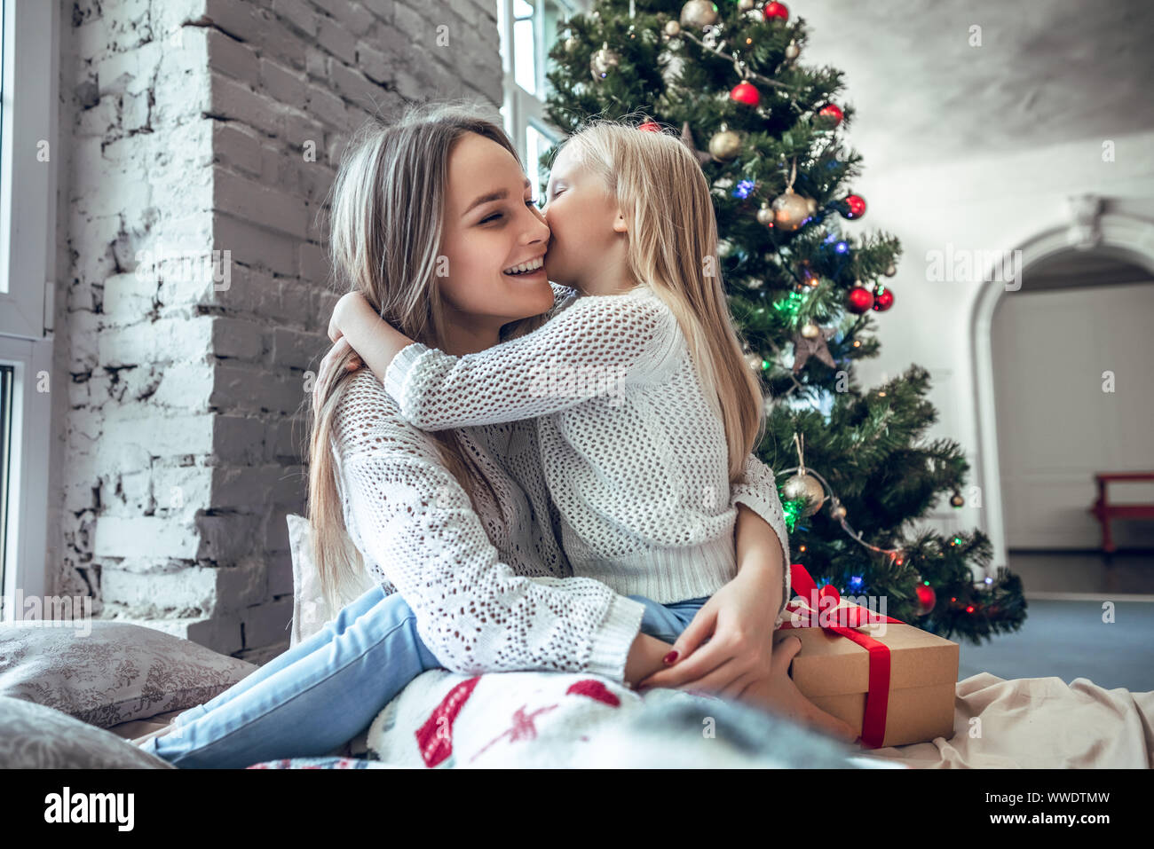 Family, holidays and happiness concept - happy daughter kissing her mother over christmas tree lights background. Winter holiday Xmas and New Year con Stock Photo