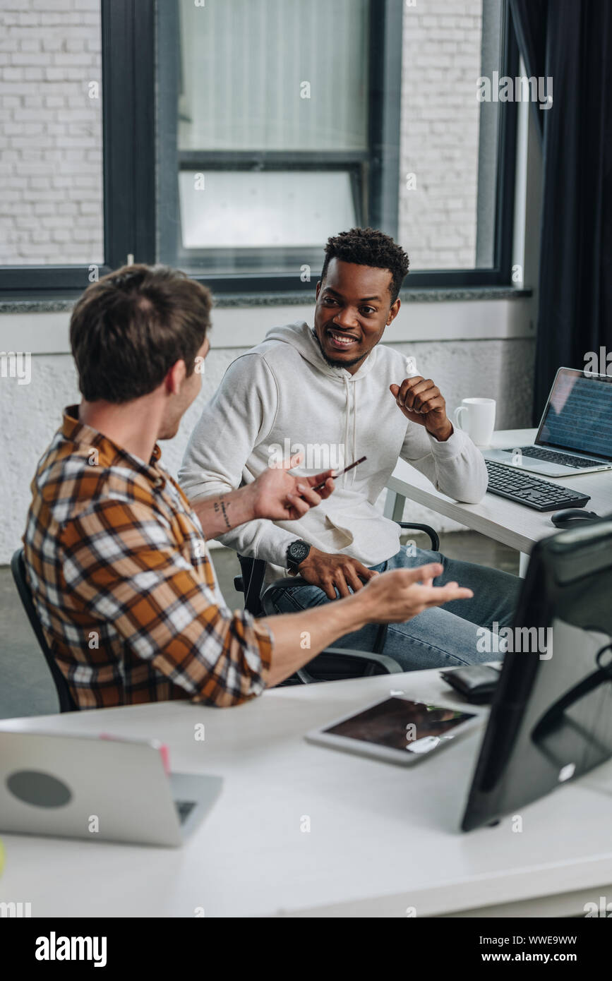 two cheerful multicultural programmers discussing ideas while sitting together in office Stock Photo
